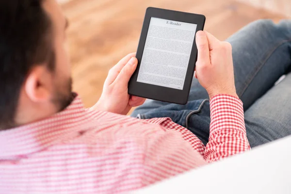 Un uomo che legge un libro in un lettore di ebook seduto su un divano a casa — Foto Stock