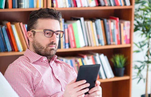 Ένας άνθρωπος διαβάζοντας ένα βιβλίο σε μια συσκευή ανάγνωσης ebook που κάθεται σε έναν καναπέ στο σπίτι — Φωτογραφία Αρχείου