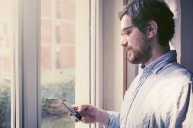 Evde, pencerenin yanında cep telefonu tutan adam.