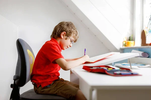 Söt liten unge pojke med glasögon hemma att göra läxor, skriva brev med färgglada pennor. — Stockfoto