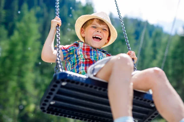 Funny kid boy having fun with chain swing on outdoor playground while being wet splashed with water — Stock Photo, Image