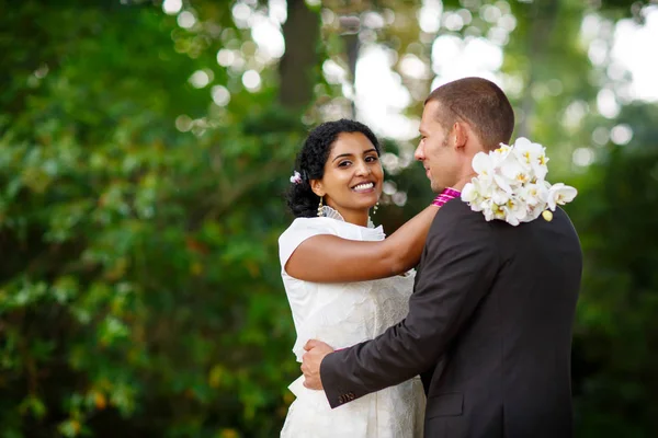 Красивая индийская невеста и кавказский жених в летнем парке. Счастливая молодая женщина с цветами. Молодой человек улыбается, пара после свадьбы — стоковое фото