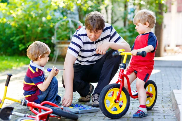 Dos niños niños felices y padre reparación de la cadena en las bicicletas y el cambio de rueda de bicicleta de equilibrio — Foto de Stock