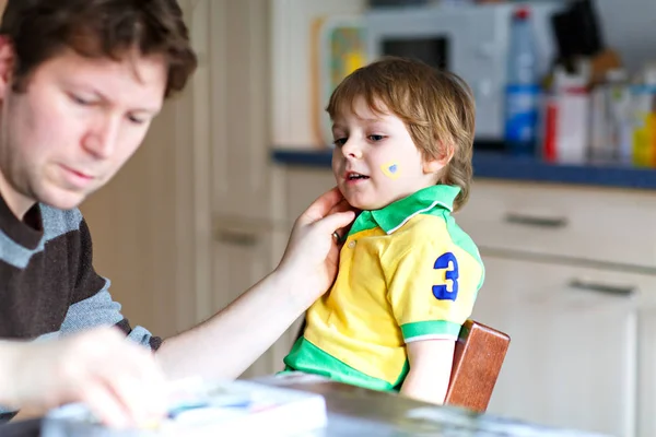 Jonge vader schilderen vlag op het gezicht van kleine zoon voor voetbal of voetbalwedstrijd. — Stockfoto