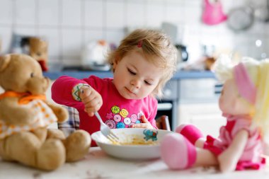 Adorable baby girl eating from fork vegetables and pasta. Little child feeding and playing with toy doll clipart