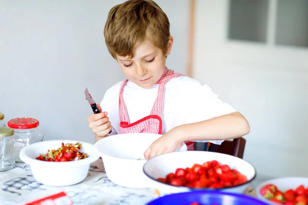 Little blond kid boy helping and making strawberry jam in summer