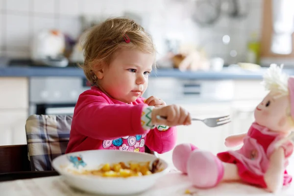 かわいい赤ちゃん女の子フォーク野菜やパスタを食べるします 餌とおもちゃの人形で遊ぶ子供 かわいい幼児 スプーンでハイチェアに座っていると ひとりでに食べ方を学ぶ娘 — ストック写真