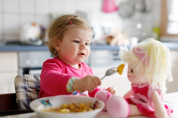 Entzückendes kleines Mädchen, das aus Gabelgemüse und Nudeln isst. Kleine Kinder füttern und spielen mit Spielzeugpuppe — Stockfoto