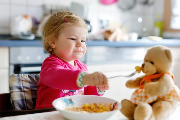 Förtjusande baby flicka äter från gaffel grönsaker och pasta. Litet barn utfodring och leker med leksak nallebjörn. — Stockfoto
