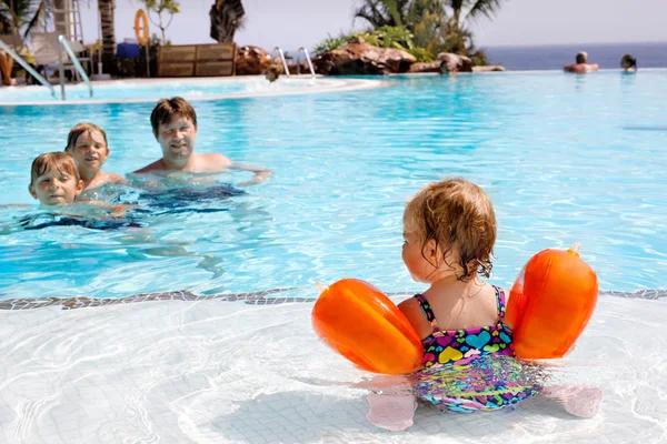 Linda niña feliz nadando en la piscina y divirtiéndose en vacaciones familiares en un complejo hotelero. Niño sano jugando en el agua. Padre y dos niños chicos, caldos, en el fondo . — Foto de Stock