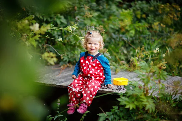 Leuk schattig peuter meisje zittend op een houten brug en kleine stenen gooien in een kreek. Grappige baby plezier met buiten spelen in de natuur. — Stockfoto