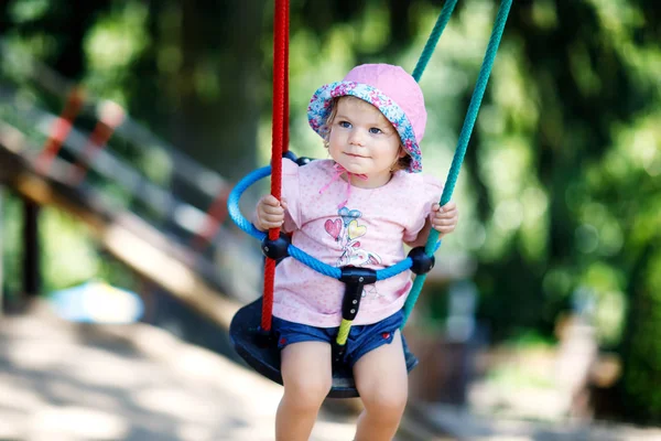 Cute adorable toddler girl swinging on outdoor playground. Happy smiling baby child sitting in chain swing. Active baby on sunny summer day outside — Stock Photo, Image