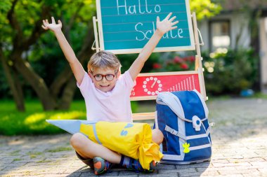 Happy little kid boy with glasses sitting by desk and backpack or satchel. Schoolkid with traditional German school bag cone called Schultuete on his first day to school clipart