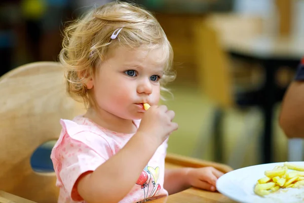 Adorable niña comiendo verduras saludables y papas fritas poco saludables. Lindo bebé feliz niño tomando comida del plato en la guardería o la cantina del vivero . — Foto de Stock