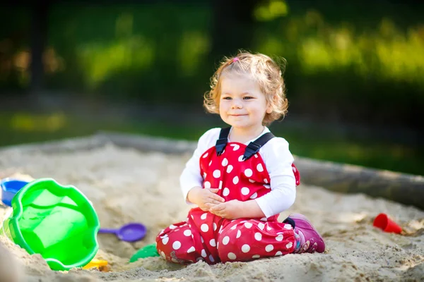 Cute toddler girl playing in sand on outdoor playground. Beautiful baby in red gum trousers having fun on sunny warm summer day. — Stock Photo, Image