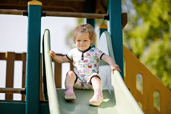 Feliz niña rubia pequeña divirtiéndose y deslizándose en el patio al aire libre. Positivo divertido bebé niño sonriendo . — Foto de Stock