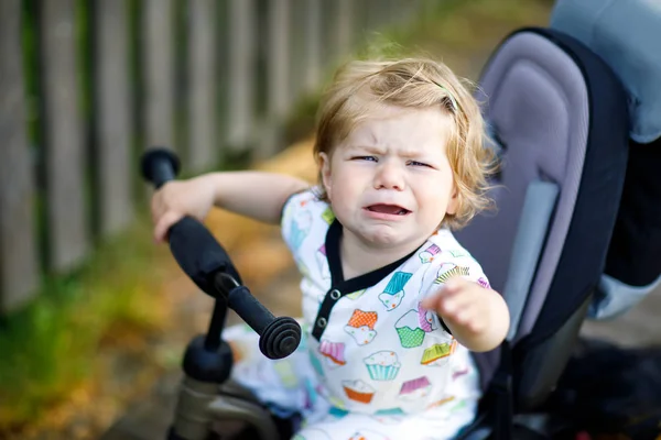 Милая очаровательная плачущая грустная девочка, сидящая на толкающем бициле или трехколесном велосипеде. Маленький ребенок собирается на прогулку с родителями в солнечный день . — стоковое фото