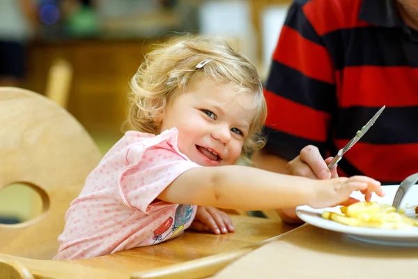 Adorable niña comiendo verduras saludables y papas fritas poco saludables. Lindo bebé feliz niño tomando comida de los padres plato en el restaurante — Foto de Stock