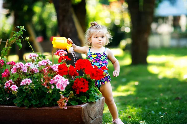 Cute little baby girl in colorful swimsuit watering plants and blossoming flowers in domestic garden on hot summer day. Adorable toddler child having fun with playing with water and can — Stock Photo, Image