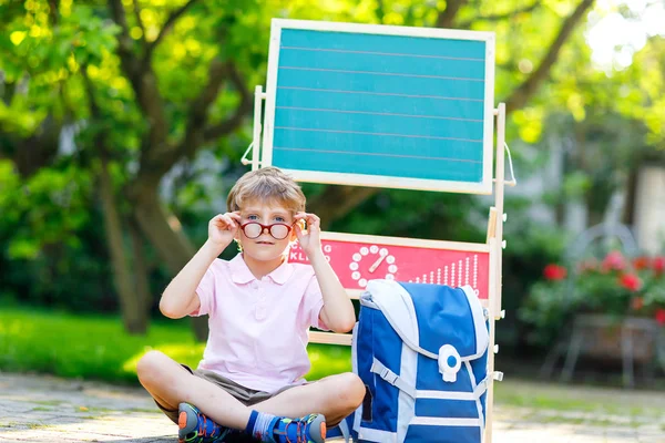 Glad liten unge pojke med glasögon som sitter vid skrivbord och ryggsäck eller axelväska — Stockfoto