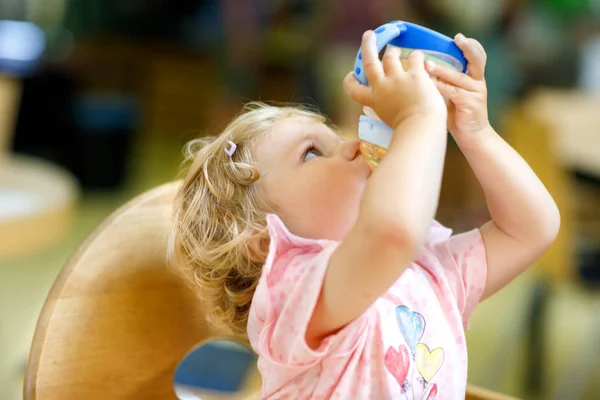 Entzückendes Kleinkind, das Formel Milch oder Wasser aus der Flasche trinkt. niedliches glückliches Baby, das Essen aus dem Teller in der Kita oder Kinderkrippe nimmt. — Stockfoto