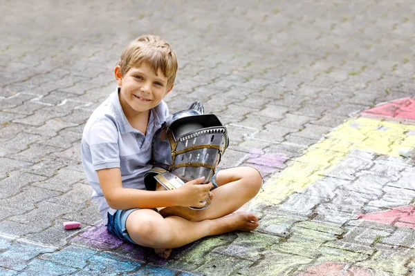 Little active kid boy drawing knight castle and fortress with colorful chalks on asphalt. Happy child with big helmet having fun with playing knight game and painting — Stock Photo, Image