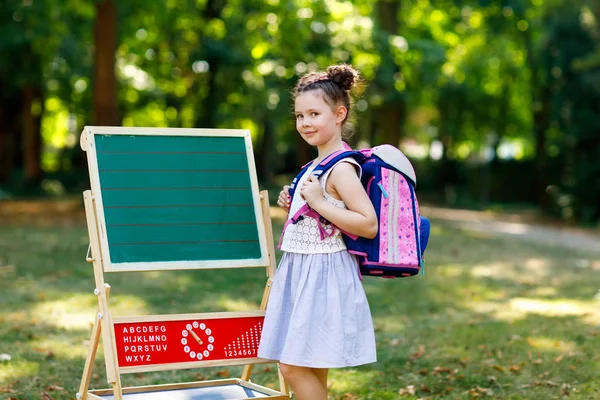 Щаслива маленька дівчинка, що стоїть за столом з рюкзаком або сателітом. Школярка в перший день початкового класу. Повернутися до концепції школи. Здорова чарівна дитина на відкритому повітрі, в зеленому парку — стокове фото