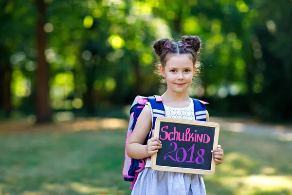 Menina criança feliz de pé com mesa e mochila ou mochila. Estudante no primeiro dia de aula elementar. Criança adorável saudável ao ar livre, no parque verde. Na mesa "Schoolchild 2018" em alemão — Fotografia de Stock