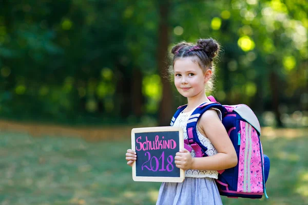 Fröhliches kleines Mädchen mit Schreibtisch und Rucksack oder Schulranzen. Schulkind am ersten Tag der Grundschule. gesundes liebenswertes Kind im Freien, im grünen Park. auf dem Schreibtisch "Schulkind 2018" — Stockfoto