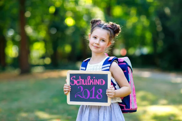 Menina criança feliz de pé com mesa e mochila ou mochila. Estudante no primeiro dia de aula elementar. Criança adorável saudável ao ar livre, no parque verde. Na mesa "Schoolchild 2018" em alemão — Fotografia de Stock