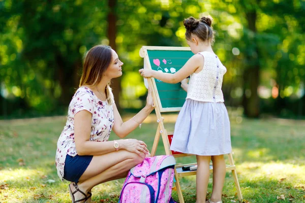 Glückliches kleines Mädchen und Mutter am großen Kreidetisch Vorschule oder Schulkind am ersten Tag der Grundschule. Zurück zum Schulkonzept. Gesundes Kind und Frau schreiben und malen im Freien — Stockfoto