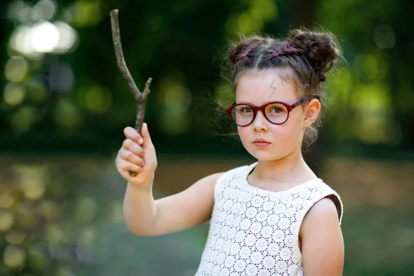 Rolig bedårande lilla kid flicka med glasögon och trä trollstav som spelar Harry Potter i parken på soliga dag. — Stockfoto
