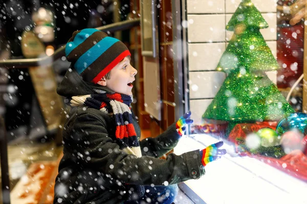 Funny happy child in fashion winter clothes making window shopping decorated with gifts, xmas tree — Stock Photo, Image