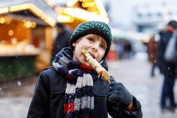 Pequeño niño lindo comiendo frutas cubiertas de chocolate blanco en pincho en el mercado tradicional alemán de Navidad . — Foto de Stock