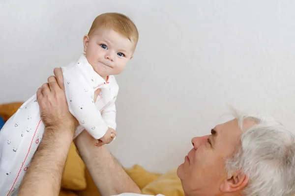 Buon nonno sano che tiene adorabile nipotina sulle braccia. Uomo anziano e bambina carina seduti, giocando e ridendo insieme a casa — Foto Stock