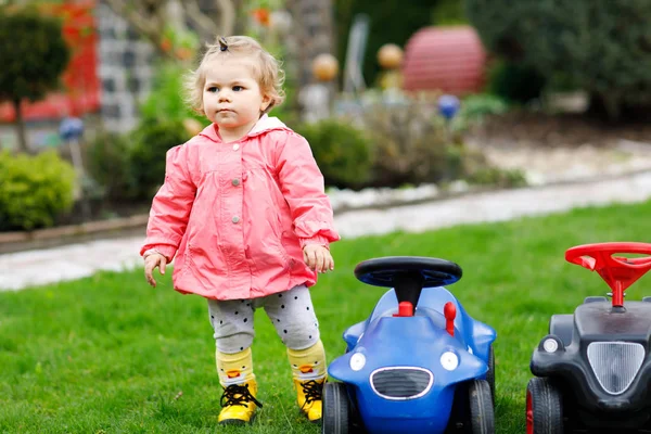 Gesunde süße kleine Mädchen spielen mit zwei Spielzeugautos im Garten. entzückendes Kleinkind, das Spaß hat. Mädchen in bunten Modekleidern. Frühling und Sommer, aktive Spiele im Freien. — Stockfoto