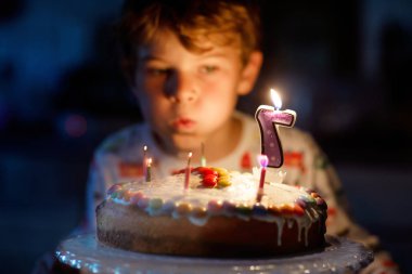 Happy blond little kid boy celebrating his birthday. Child blowing seven candles on homemade baked cake, indoor. Birthday party for school children, celebration of 7 years clipart
