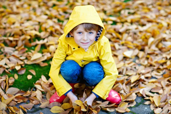 Portrait of happy cute little kid boy in yellow rain coat and red rubber boots with autumn leaves background. Funny child having fun and playing in fall forest or park on cold autumnal day — Stock Photo, Image