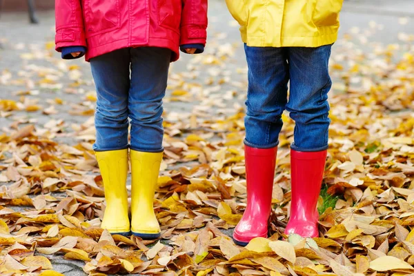 Duas crianças brincando em botas de borracha vermelha e amarela no parque de outono em casacos de chuva coloridos e roupas. Fechar-se de pernas de crianças em sapatos dançando e andando através de folhas outonais queda e folhagem — Fotografia de Stock
