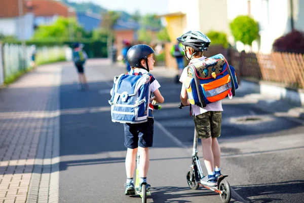 Due scolaretti in casco di sicurezza a cavallo con scooter in città con zaino nella giornata di sole. Bambini felici in vestiti colorati in bicicletta sulla strada per la scuola. — Foto Stock