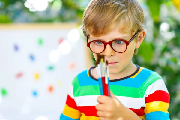 Confused little kid boy with glasses holding watercolors and brushes. Happy child and student is back to school. Education, school, learning concept. School, preschool nursery equipment. — Stock Photo, Image