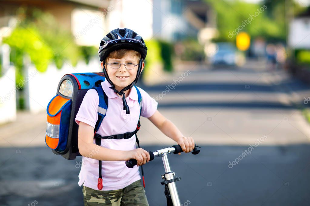 Active school kid boy in safety helmet riding with his scooter in the city with backpack on sunny day. Happy child in colorful clothes biking on way to school.