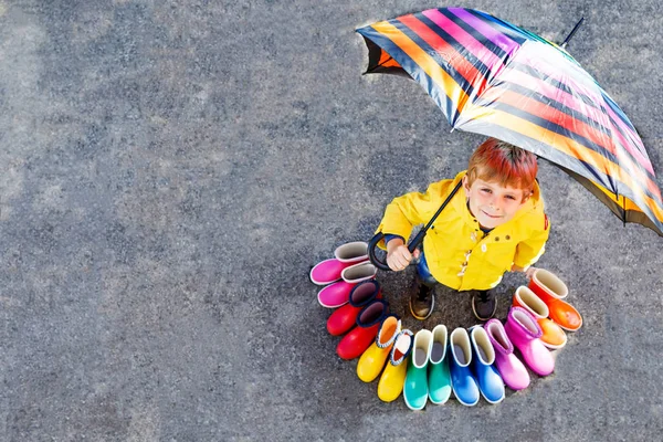 Ragazzino e gruppo di stivali da pioggia colorati. Bambino biondo sotto l'ombrello. Primo piano di scolaro e stivali di gomma diversi da angolo alto. Calzature per caduta piovosa — Foto Stock