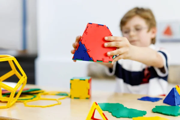 Маленький мальчик в очках играет с пластмассовым набором элементов в школе или детском саду. Счастливый ребенок строит и создает геометрические фигуры, учится игре и геометрии: — стоковое фото