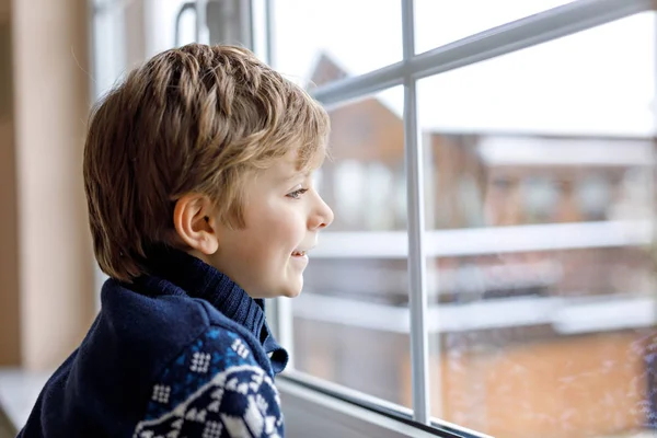 Šťastný rozkošný chlapeček sedí u okna a dívá se ven na sníh na Štědrý den nebo ráno. Usmívající se dítě fascinované sněhem a velkými sněhovými vločkami — Stock fotografie