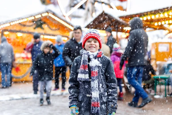 Little cute kid boy having fun on traditional German Christmas market during strong snowfall.. Happy child enjoying traditional family market in Germany, Munich. Laughing boy in colorful clothes — Stock Photo, Image