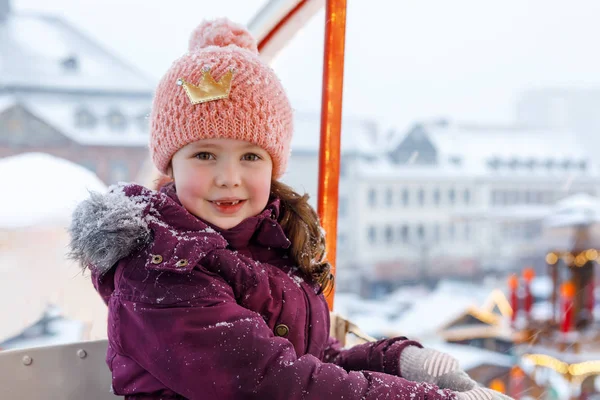 Little cute kid girl having fun on ferris wheel on traditional German Christmas market during strong snowfall. Happy child enjoying family market in Germany, Munich. Laughing girl on carousel: — Stock Photo, Image