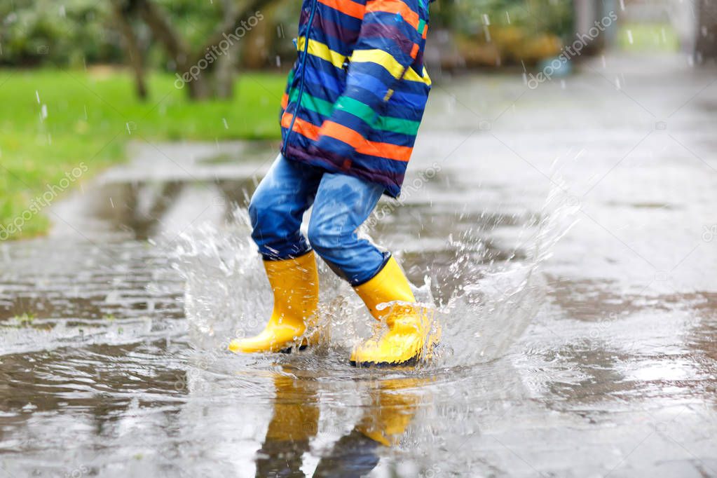 Close-up of kid wearing yellow rain boots and walking during sleet, rain and snow on cold day. Child in colorful fashion casual clothes jumping in a puddle. Having fun outdoors