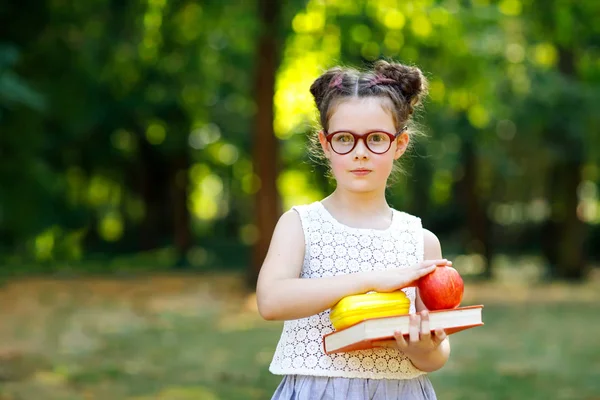 Funny adorable little kid girl with glasses, book, apple and backpack on first day to school or nursery. Child outdoors on warm sunny day, Back to school concept. Healthy child of elementary class.