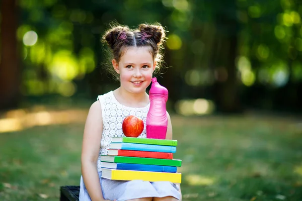 Szczęśliwy adorable kid dziewczynka czytanie książki i gospodarstwa różnych kolorowych książek, jabłka i butelka wody na pierwszego dnia do szkoły lub przedszkola. Powrót do koncepcji szkoły. Zdrowe dziecko elementarne klasy. — Zdjęcie stockowe